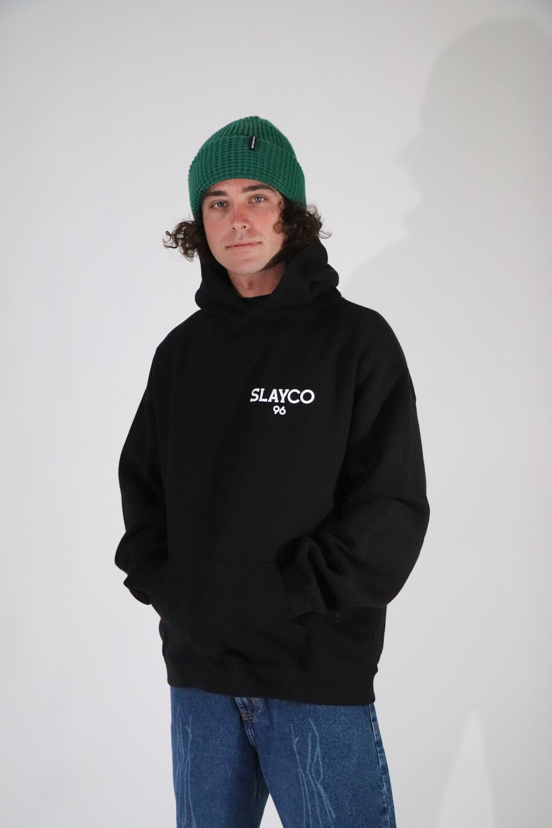 Axell Hodges wearing a black hoodie with  slayco text and a green beanie on a light gray background